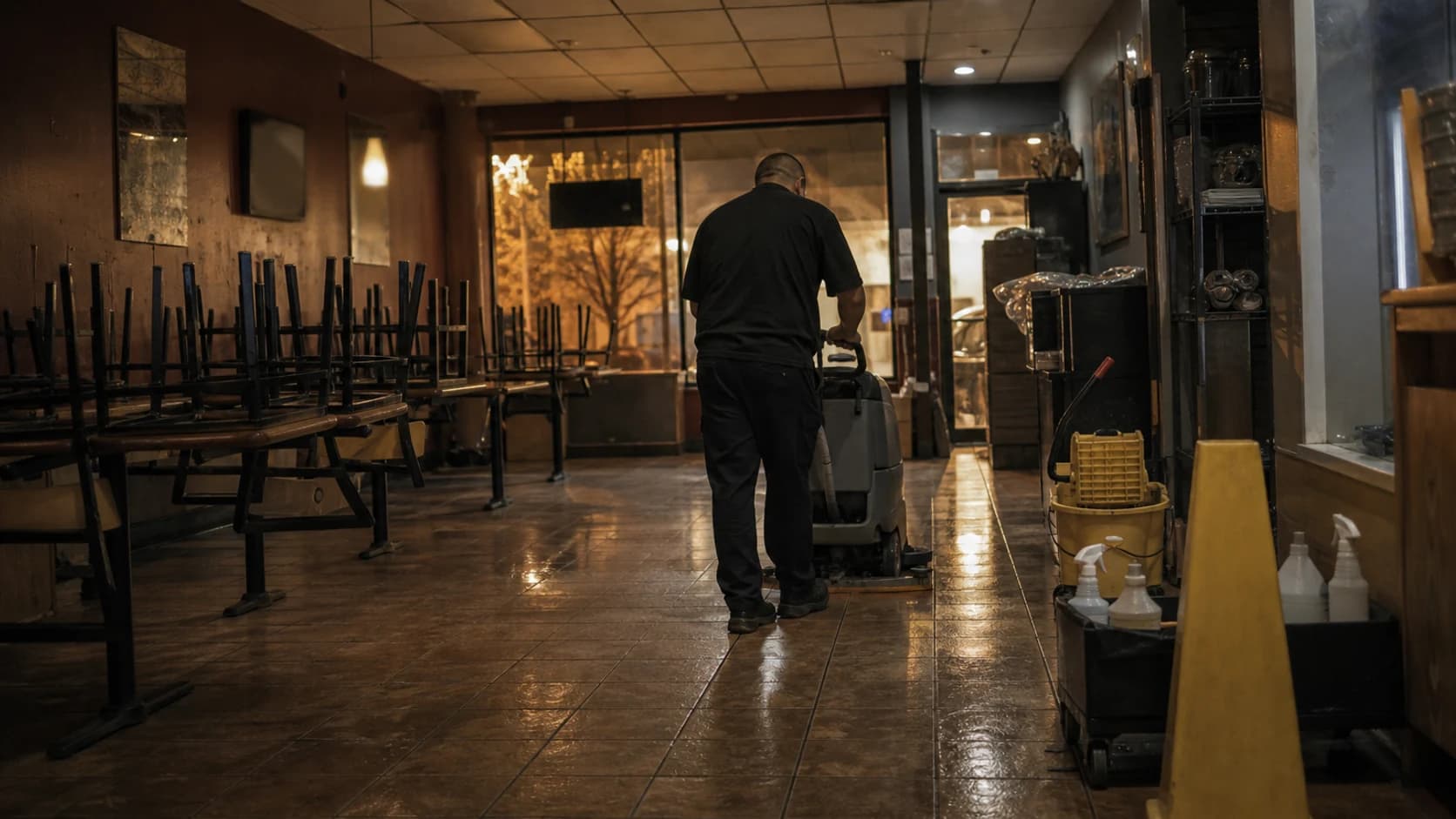 A cleaner running a floor scrubber through a restaurant dining room after closing